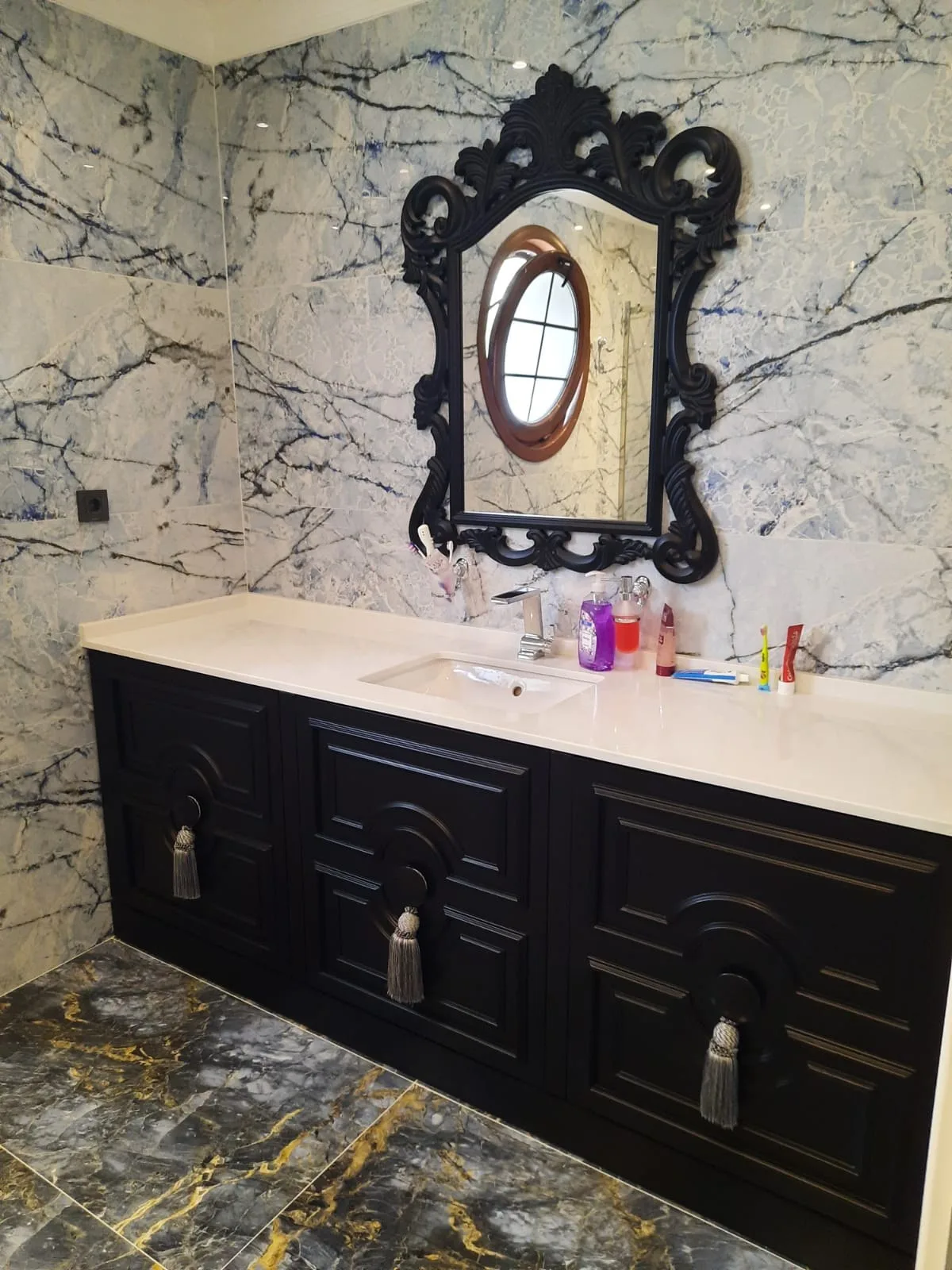 Siyah oymalı panel kapaklı banyo dolabı; püskül kulplar ve mavi damarlı mermer duvar